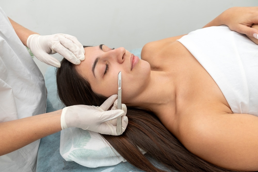 Woman laying on exam table and receiving a dermaplaning treatment on her face.