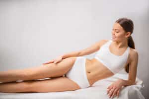Young woman in a white bikini smiles as she touches her smooth legs, feeling glad she skipped waxing before laser hair removal
